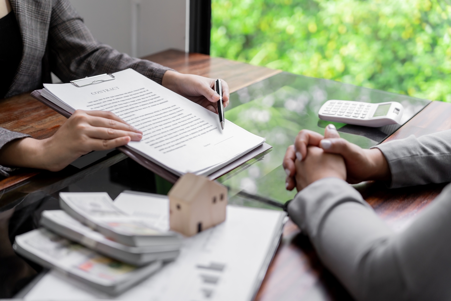 Beratungsgespräch zum Hausbau: Person erklärt Kosten anhand von Dokumenten, Taschenrechner und Hausmodell auf dem Tisch